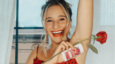 Woman smiling with blonde hair in a bun wearing a red and black polka dot tank top lifting her arm up and holding a rose and a Rose+Black Pepper Hemp Seed Oil deodorant from Schmidt's